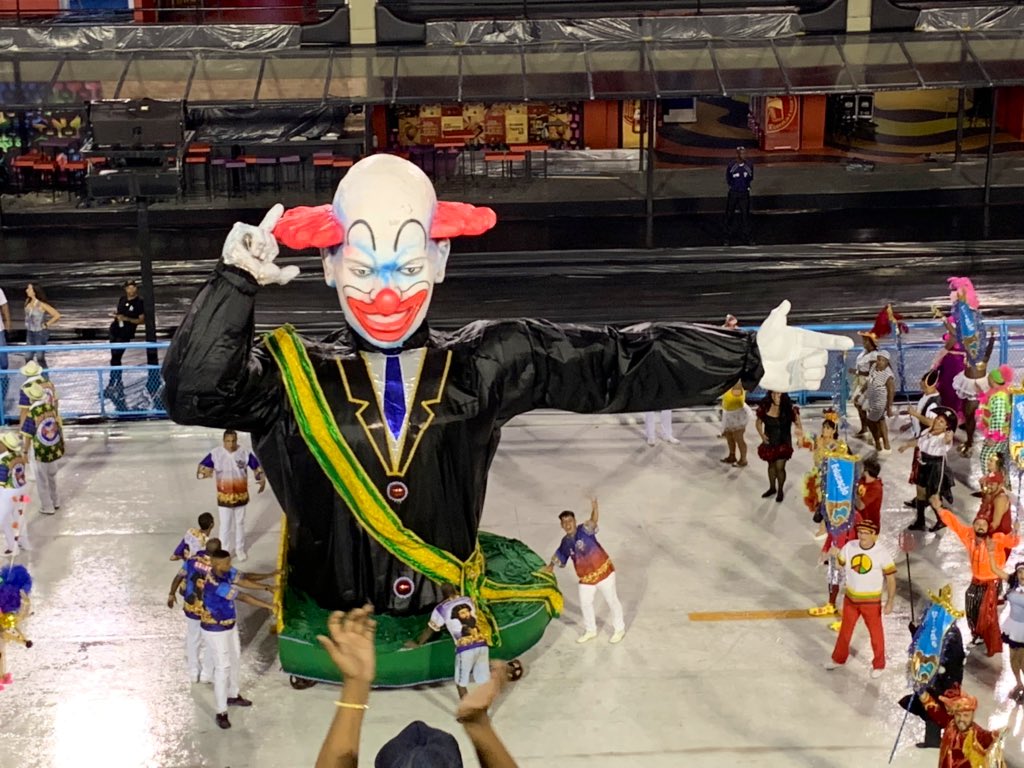 Carnavalize's tweet photo. Elemento cenográfico com faixa presidencial e cara de palhaço fecha o desfile da escola, apontando “arminha” de dedo para um bloco de foliões segurando estandartes, que pediam: edução, saúde, cultura e outros desejos. #Vigario 🔵 #CarnavalizeRJ 🔵 https://t.co/bS9KUycdpT