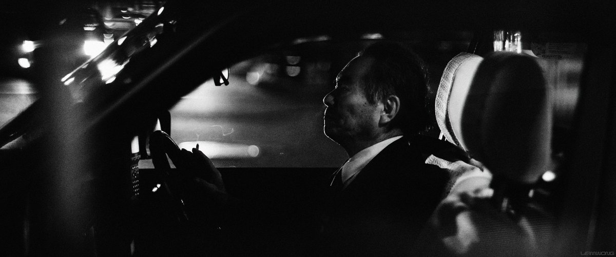 Night time photography by Liam Wong. Black and white. A taxi driver side-on view whilst he is in his cab. He's smoking a cigarette - his hand is near the steering wheel, smoke is coming from the lit cigarette.