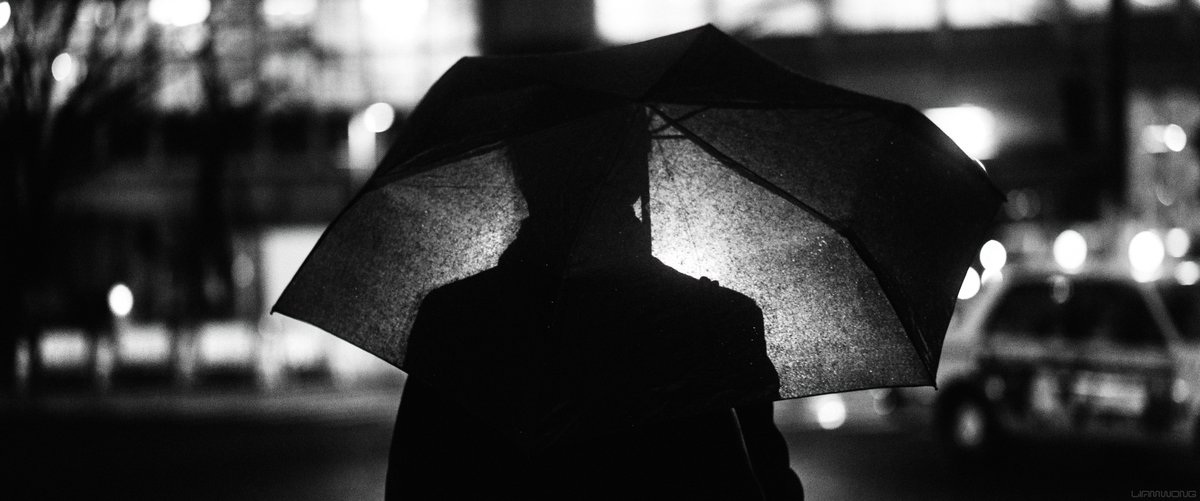 Night time photography by Liam Wong. Black and white. A silhouette of a man with an umbrella. His silhouette is lit up by the traffic in front. His figure can be seen through his black umbrella. It is a medium shot from the torso upwards. He is framed centrally.