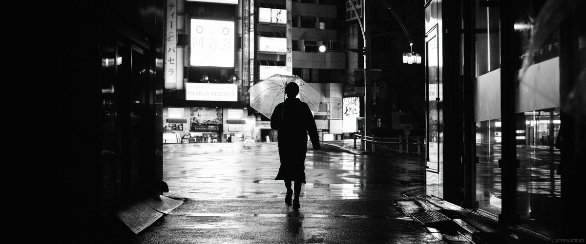 Night time photography by Liam Wong. Black and white. A silhouette of a man with a transparent umbrella. His silhouette is lit up by the lights in front reflecting off the street. It is a full body shot, he is composed centrally.