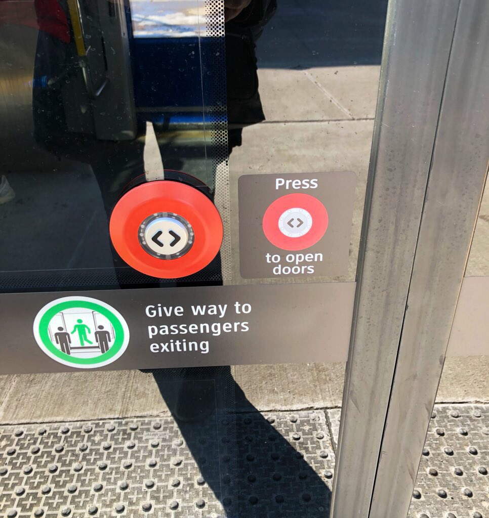 A photo of a tram door with a big red button with angle brackets <> on it to indicate that it opens the door. Next to the button is a sticker with an image of the button and the words “Press to open doors”. There’s also a sticker that says “Give way to passengers exiting”