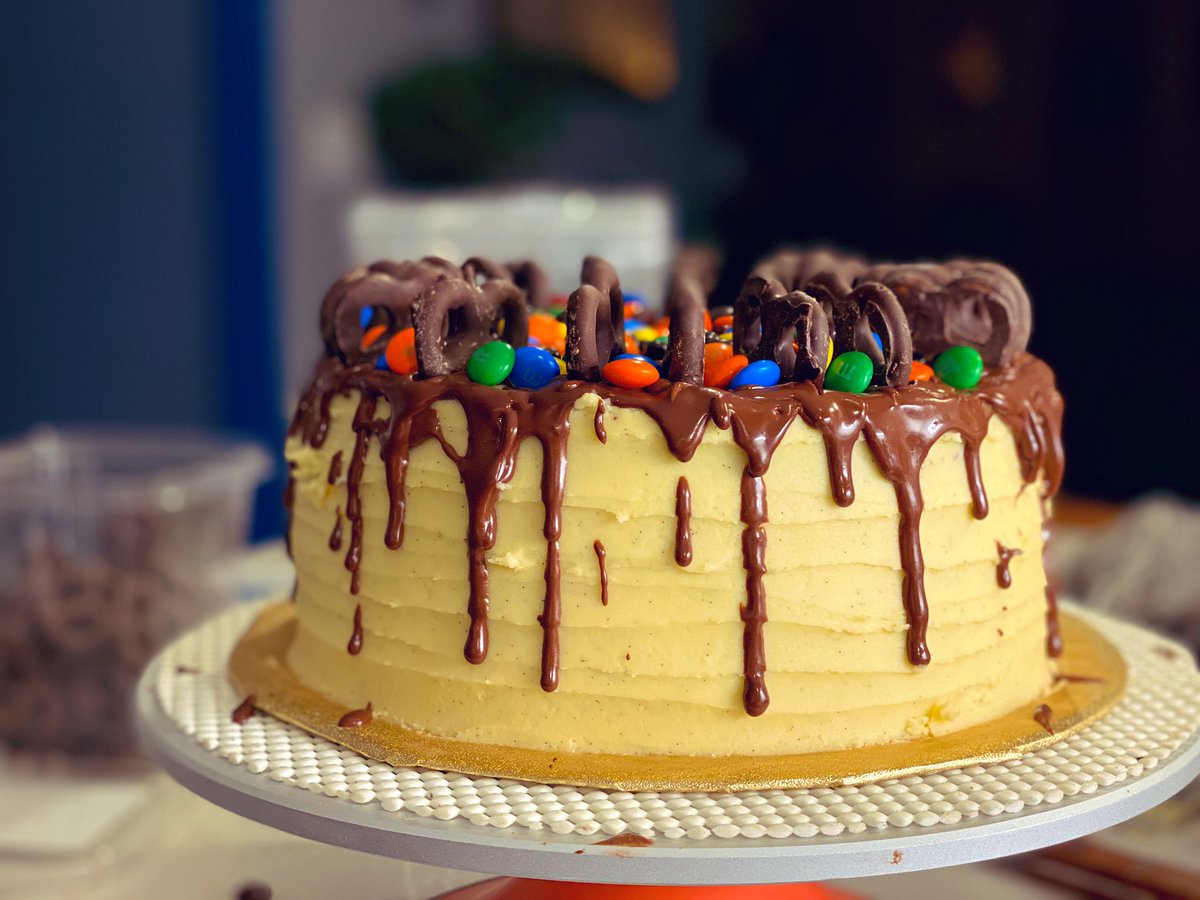 Cake on a stand covered with vanilla frosting, chocolate dripping down the sides, and topped with chocolate candies and chocolate-covered pretzels.