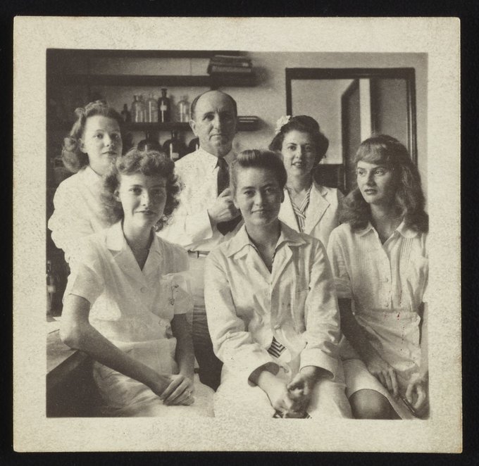 Sepia-toned photo of 5 women and 1 man in a lab. They are all wearing white lab coats.