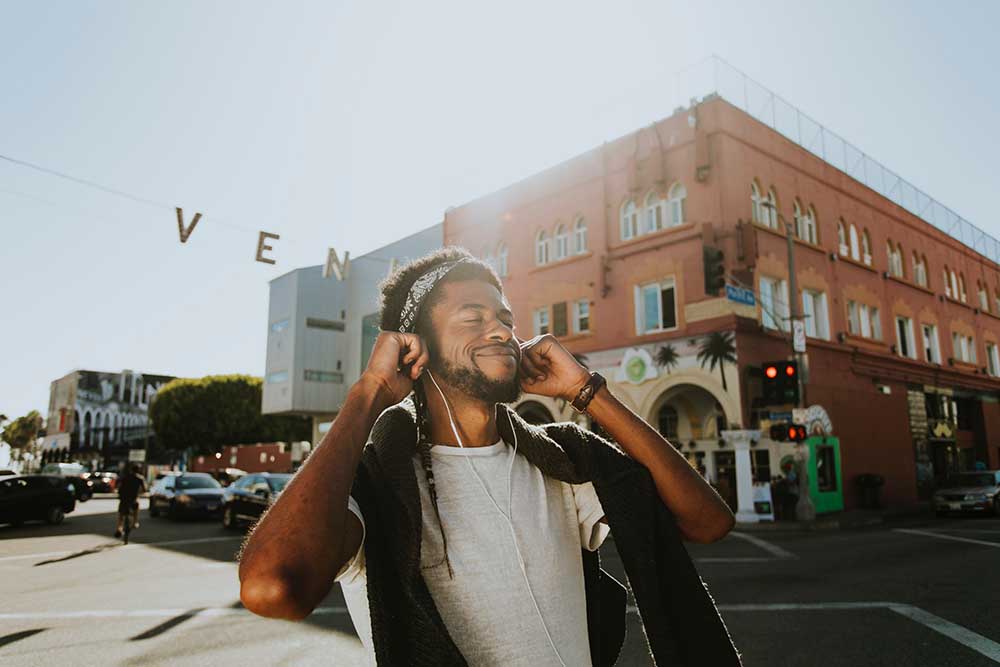 a young Black man smiling with eyes closed listening to headphones outside on a sunny day