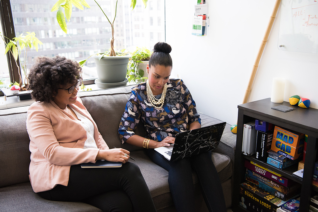 two women sitting down together on a couch sharing a laptop smiling looking at the screen