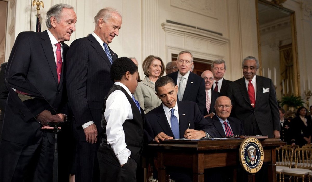 Photograph of President Barack Obama signing the Affordable Care Act surrounded by lawmakers and VP Joe Biden. A young boy is standing next to Obama as he signs. 