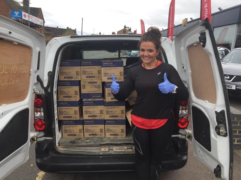 A woman is standing outside. She is giving the thumbs-up sign. She is in front of a van. The van has its back doors open. There are boxes in the back of the van.