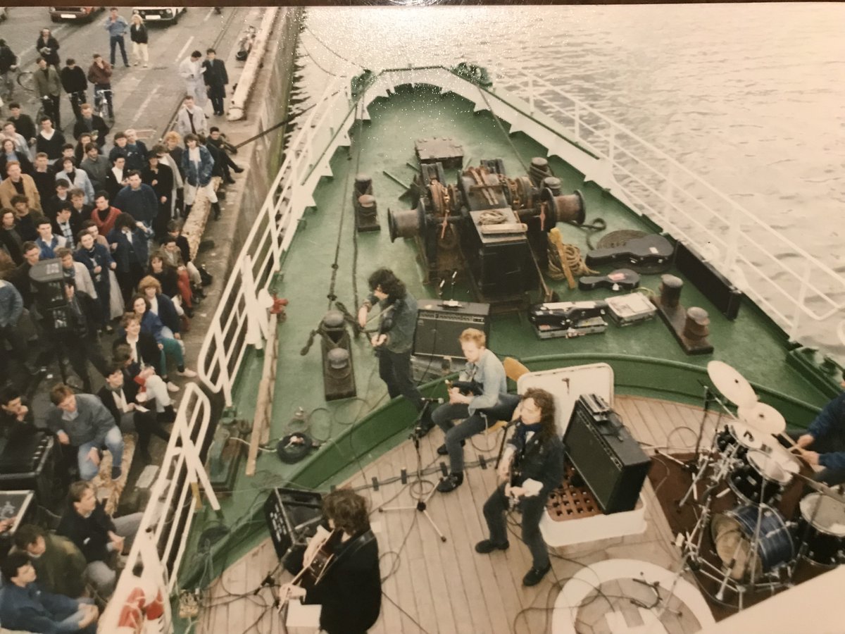 MickPuck's tweet photo. Odd moments in #TheWaterboys history: show on deck of #Greenpeace ship, Sirius, May 1987, Dublin docks. I like how #SteveWickham has feet in different worlds. #VinnieKilduff in denim playing uillean pipes. #FranBreen, now a band member, on drums. https://t.co/kjCgwoHUT8