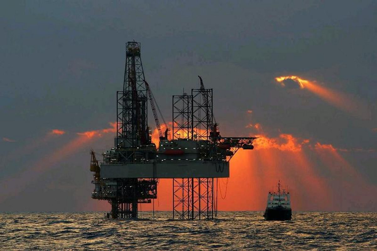 A large boat drives by a massive oil & gas platform rising above the ocean on tall metal stilts under a colorful sunset sky.