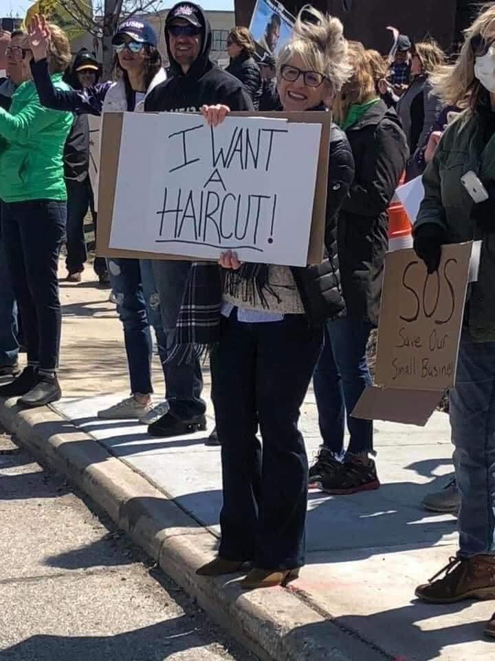 White woman surrounded by other white people. Holds sign reading “I need a haircut.”