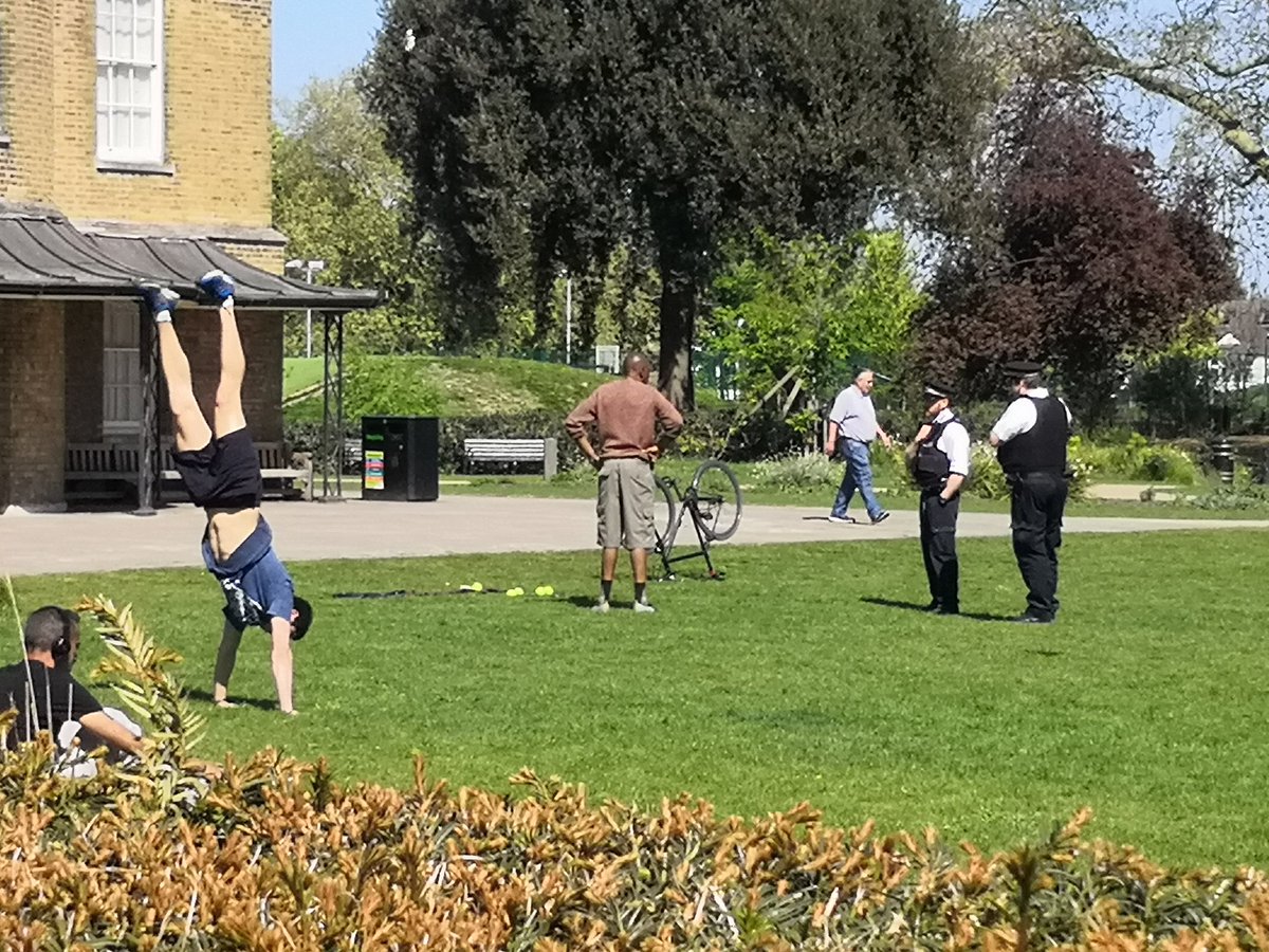 Black guy being told to leave Clissold Park by 2 police officers while white guy practises handstands nearby.