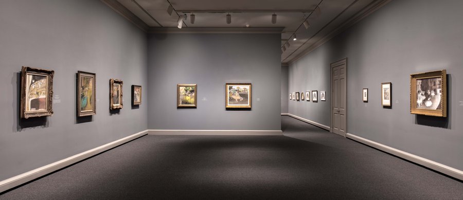 An installation view of a gallery space with paintings hung on three walls and another gallery seen in the distance.
