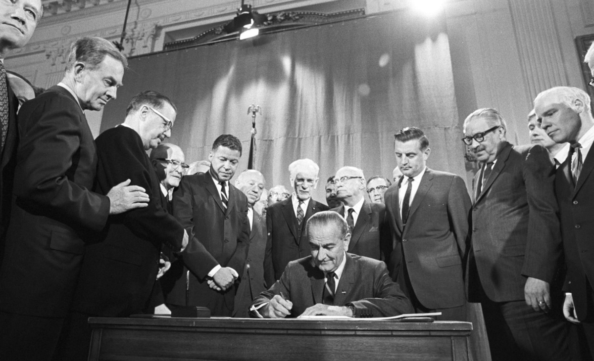 A photo from 1968 of President Johnson sitting at a desk and signing the Fair Housing Act, with lots of men standing around him to watch.