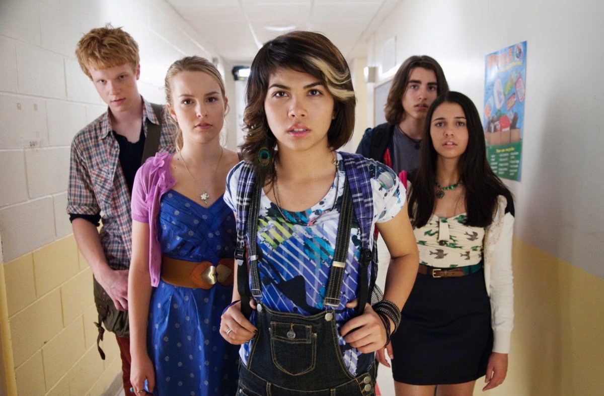 Image is from the film Lemonade Mouth (2009). Five students - three girls and two boys - standing in all school corridor look at a something placed on the wall in front of them.