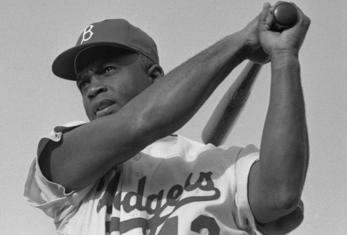 A photograph of Jackie Robinson swinging his bat and wearing a baseball cap and Dodgers uniform.
