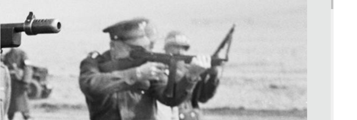 In the background you can see Supreme Allied Commander General Eisenhower shooting the later simplified M1 version of the Thompson, with a long 30-round magazine.