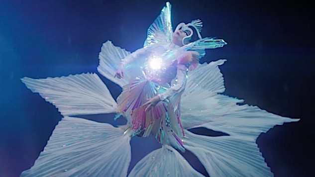 The Gate (2017)The video begins with Björk playing the flute on a cliff's edge, while several objects levitates. The 2nd part of the video portrays Björk passing a glowing orb to an "all-crystal being". A glow in her chest projects 3 other figures, that start dancing around her