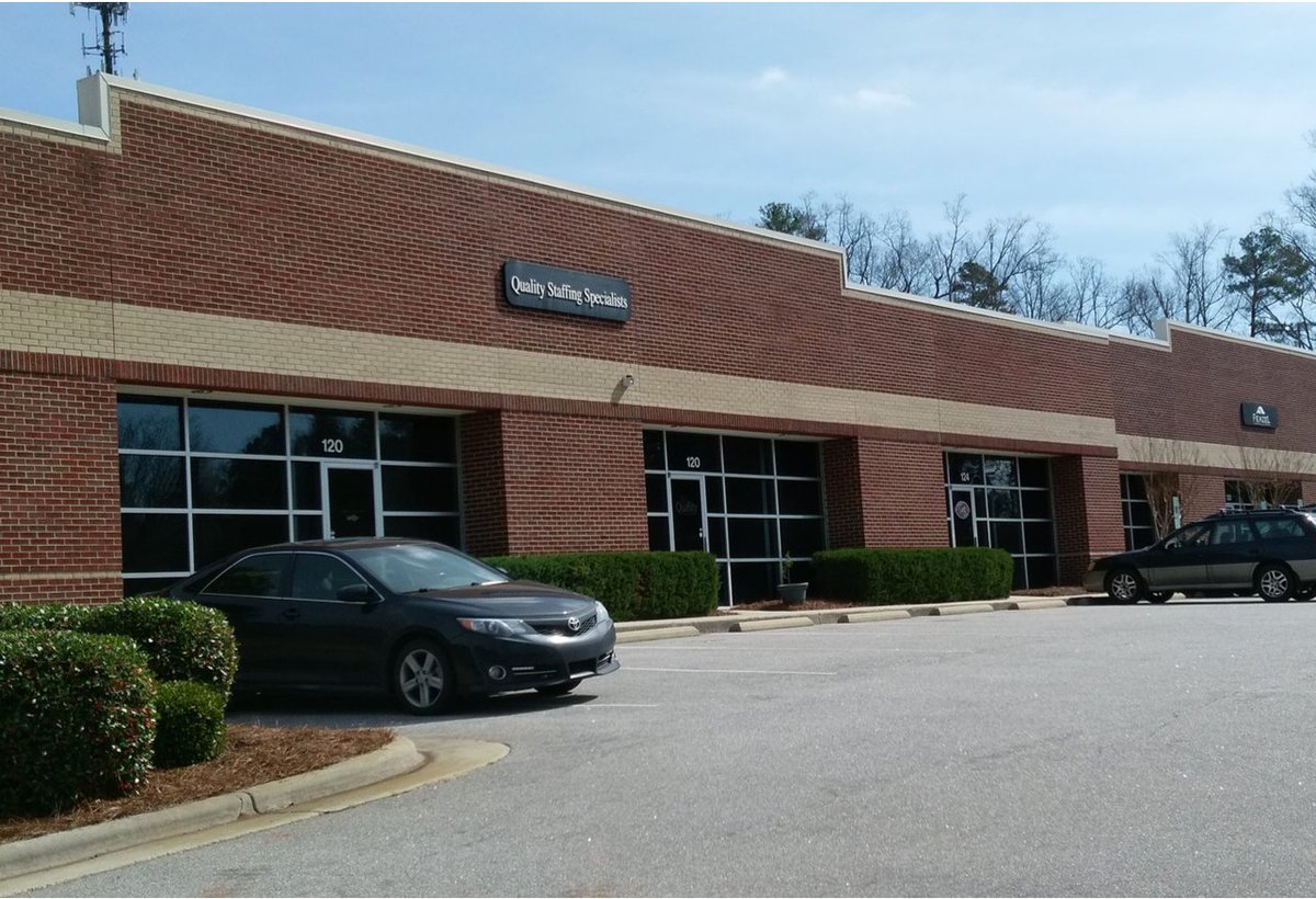 A photo of a brick office building shot from the parking lot. A single vehicle is parked on the left in front of the building and next to a small landscaping strip with grass and hedges. The business is called "Quality Staffing Specialists." There are small landscaping strips in front of the building planted with hedges. There is a cellular mast visible in the background on the left; on the right, trees are visible.