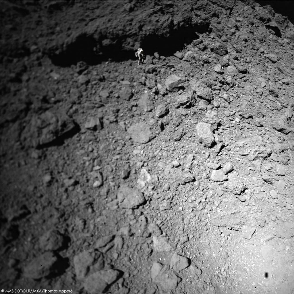 MASCOT a pris cette 1ère photo depuis une altitude de 27,5 m. En bas à droite, l'ombre de l'atterrisseur, long de 30 cm, donne l'échelle. Le gros rocher en haut de l'image est celui qu'on voyait à droite dans l'animation précédente. L'astronaute Harrison Schmitt donne l'échelle.