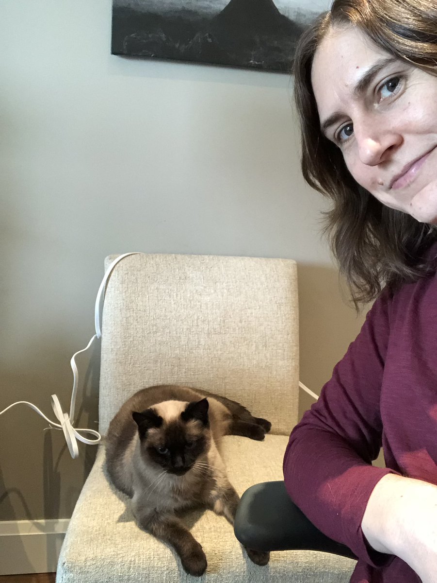 Me leaning to the side in an office chair, smiling next to a cushioned chair on which the cat is sitting, looking very serious.