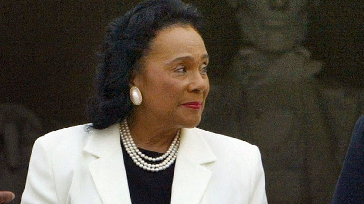 Photograph of Coretta Scott King looking to the right. She is wearing large earrings and a pearl necklace. Visible behind her is the Lincoln Memorial.