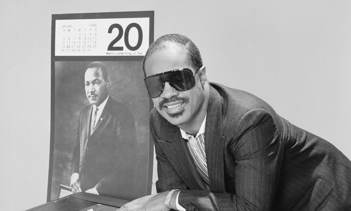 A photo of Stevie Wonder standing next to an image of Dr. Martin Luther King, Jr., which shows a calendar that highlights MLK Day. Stevie Wonder had a role in the holiday's creation.