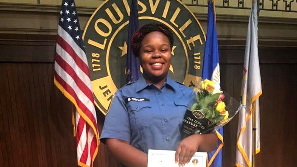 A photo of Breonna Taylor in her uniform, holding a bouquet of flowers, standing in front of a Louisville seal and flags, including the American flag.