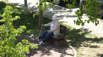 Yaşlı bir kadın parkta bir bankta tek başına oturuyor. Sağ elinde bastonu kafasında beyaz bir şapka var.