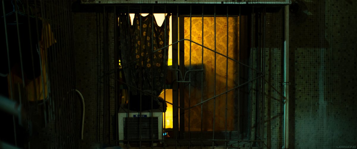 Photography by Liam Wong of Chongqing, China at night. An isolated close-up of a window. It is yellow in color. It feels old and worn. The exterior is made up of small tiles. There is a curtain with flowers. A man is seen on his phone through the small gap in the window. It is mostly green and warm tones.