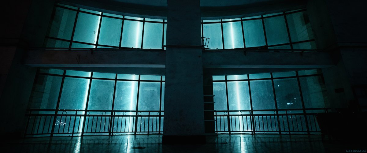Photography by Liam Wong of Chongqing, China at night. An empty residential window looking out. Several neon strips of light illuminate through the windows. The structure feels like a cross. It is a desaturated blue color.