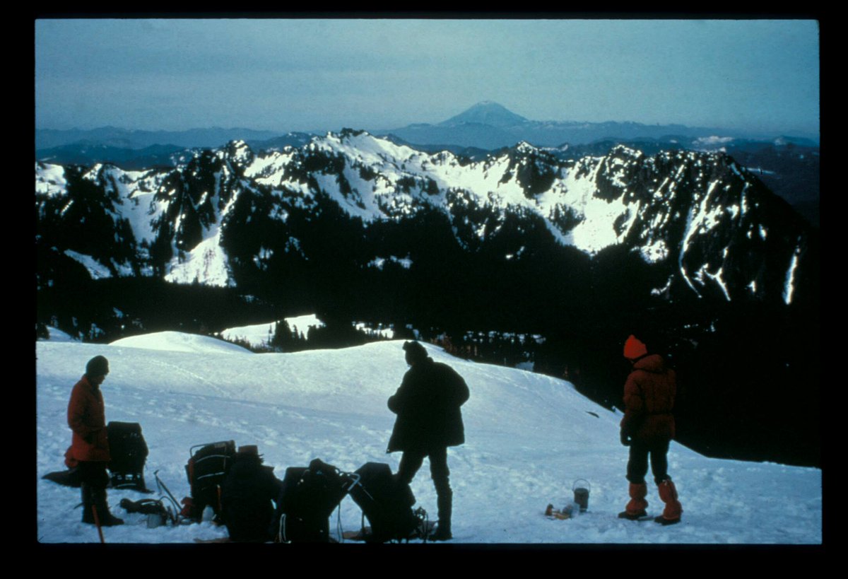 Hikers on a snowy slope look over mountain ridges towards a distant volcanic peak. 