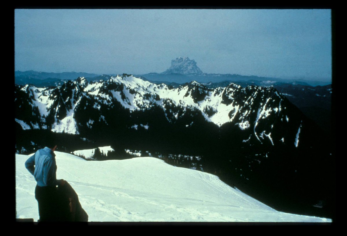 A hiker observes the start of an explosion from a distant volcanic peak.