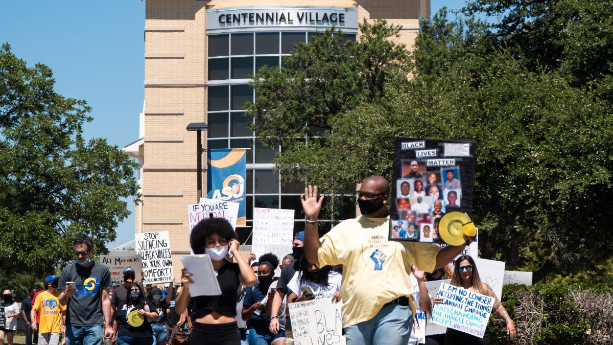 Group of people walking down the Angelo State mall holding signs with Centennial Village behind them.