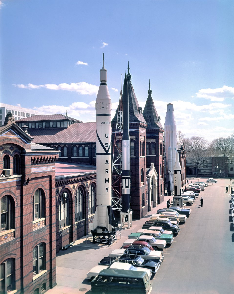 Color photograph of cars parked outside of the Arts and Industries Building where rockets are displayed. 