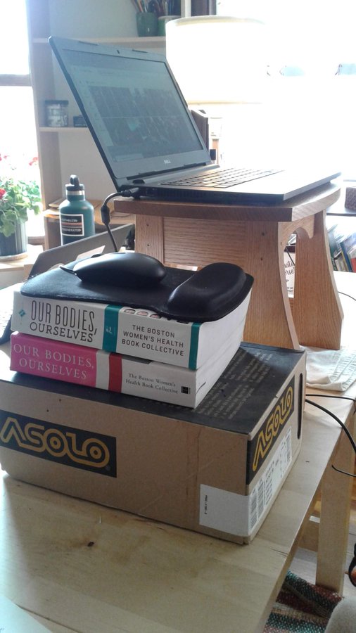A laptop sits atop a footstool on a table with a mouse and mousepad beside it on a shoebox and pile of books.