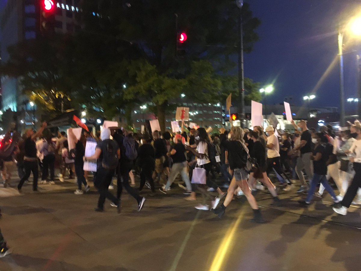 Cars on Main are joining in the chant of Black Lives Matter. The crowd is assembled behind a flag that says Black Lives Matter. The protest is moving along Main and has crossed Peabody, to the area of N Main that is restricted to trolly service.