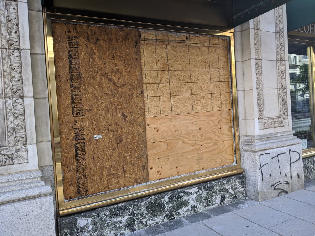 Busted out window is boarded up at Geoffrey Lewis tailors. Other window has graffiti.