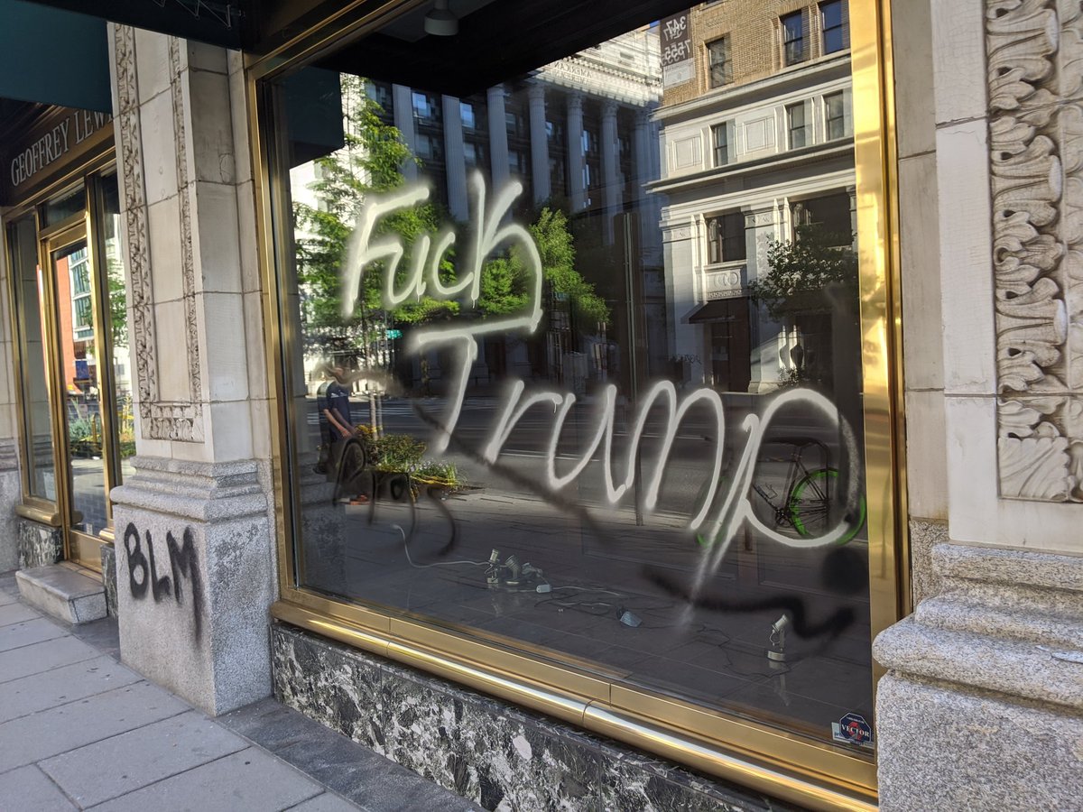 Busted out window is boarded up at Geoffrey Lewis tailors. Other window has graffiti.