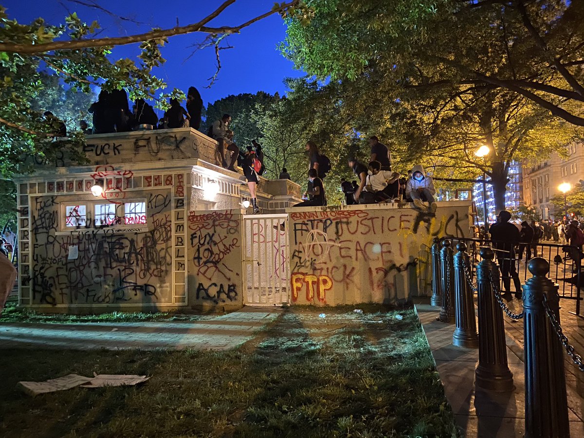 A group of protesters are sitting above a small graffitied building in the park. At least some appear to be hurling water bottles at the police from there – at  Lafayette Square Park