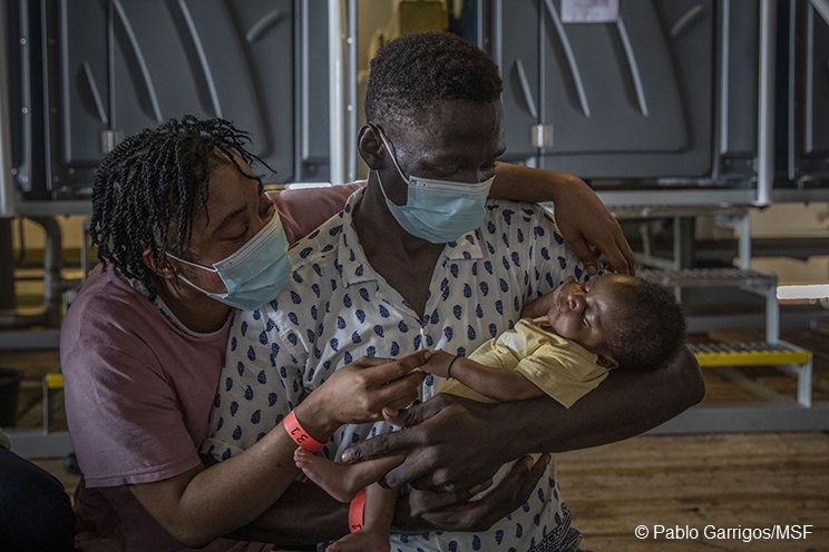 Ein Mann hält ein baby im Arm und schaut es an, er hat eine Gesichtsmaske auf. Eine Frau steht hinter ihm und hat einen Arm um ihn gelegt, sie schaut ebenfalls das Baby an und hält die Hand des Babys. Sie hat auch eine Maske an.