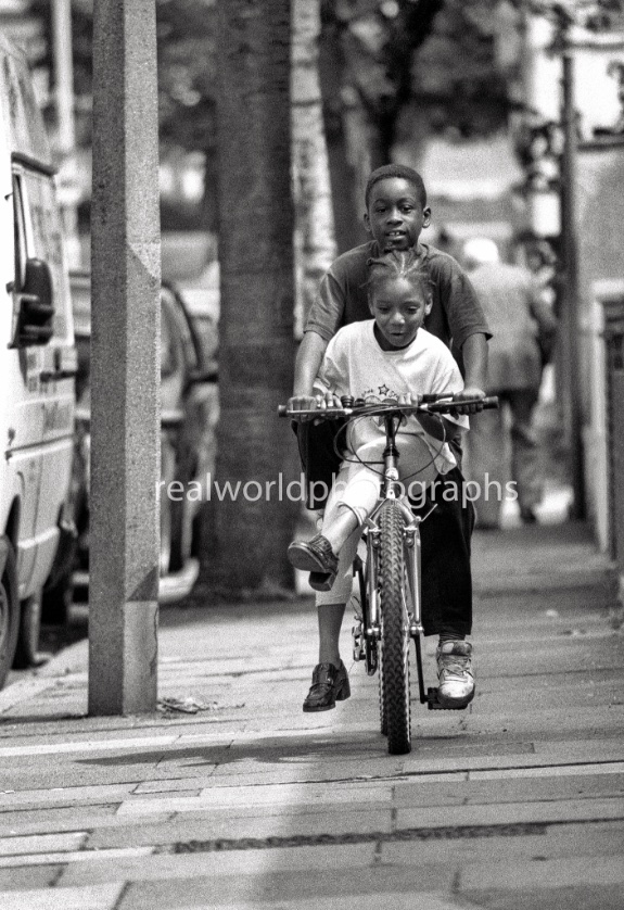 A sibling gets a double-ride in Wood Green, north London, England. Gary Moore photo. Real World Photographs. #photography #london #photojournalism #woodgreen #ldn #uk #realworldphotographs #people #children #images #blackandwhite #malmo #sweden #streetphotography #street 