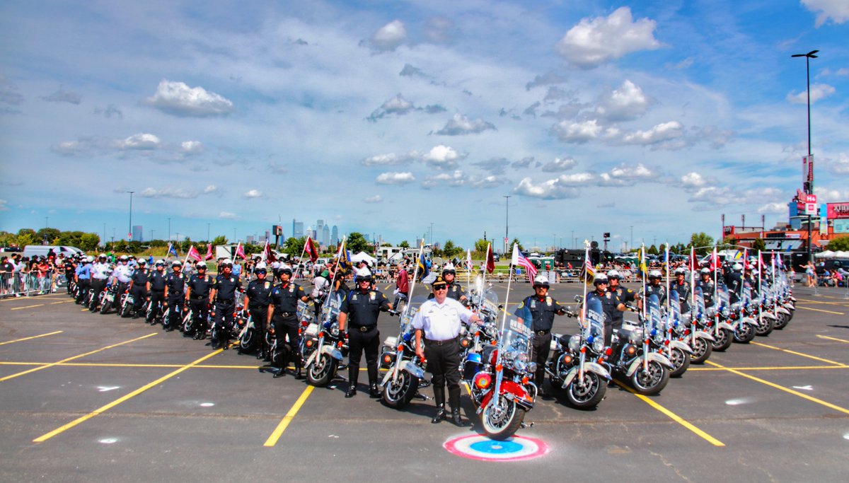 about two dozen police officers on motorcycle drill team stand in V formation next to their motorcycles with flags attached in large parking lot in daylight 