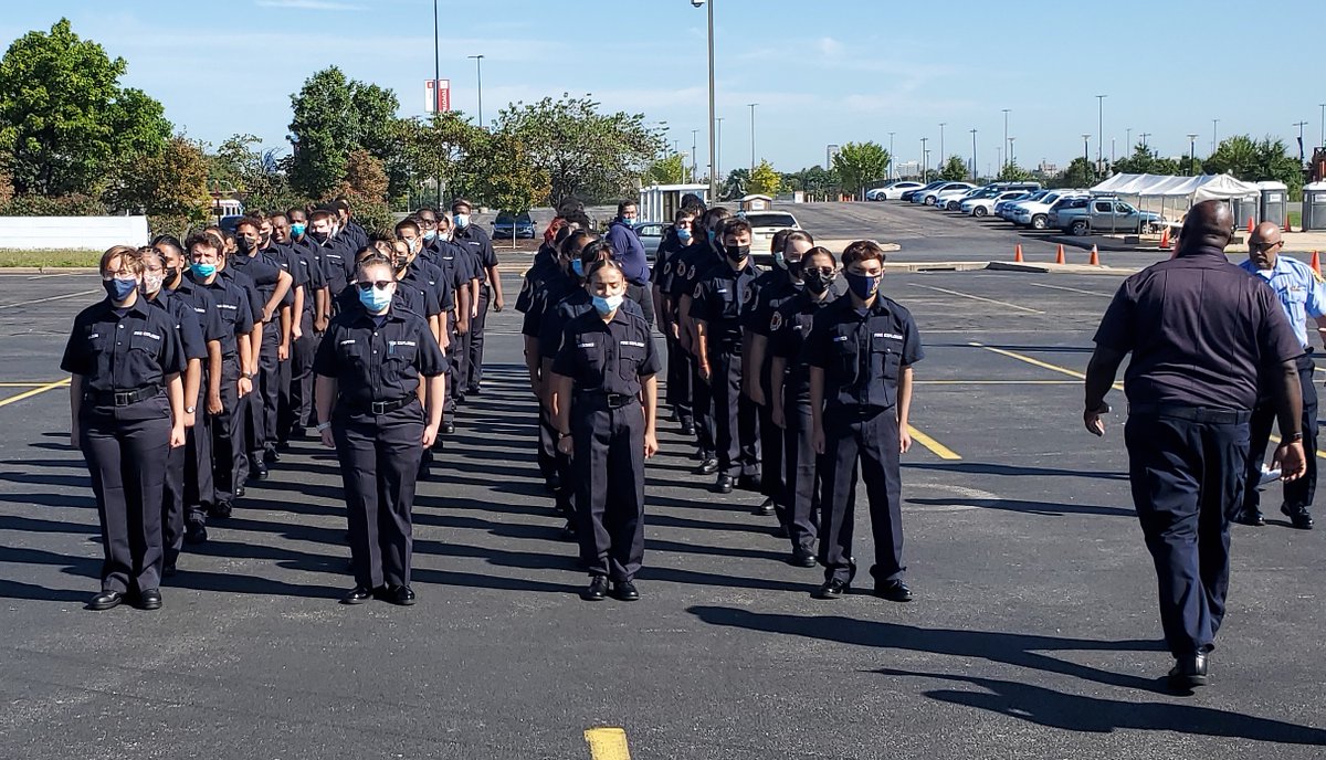 about three dozen teenagers in uniform and face masks standing in four lines in formation in parking lot in daylight