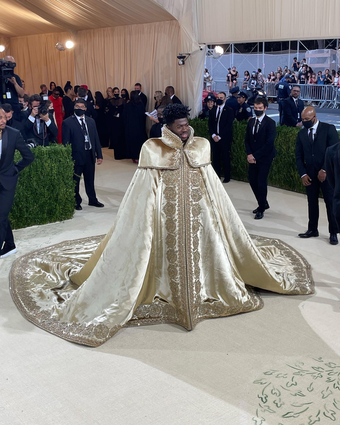 American rapper Lil Nas X stands on The Met’s red carpet. He is a dark-skin-toned young man wearing a long, elaborate gold cape, like a royal mantle, with embroidery along its edges and prominent shoulders. Security guards stand in the background.