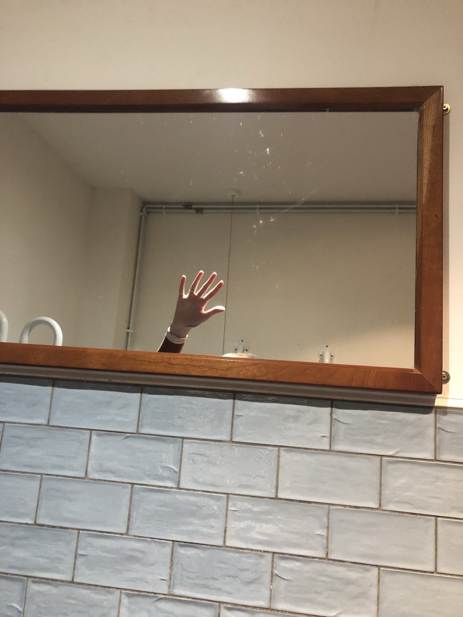 A mirror hanging on a bathroom wall. You can see a hand waving in the mirror, demonstrating that the mirror is hung at a height that is too high up and above the eye level of a wheelchair user.