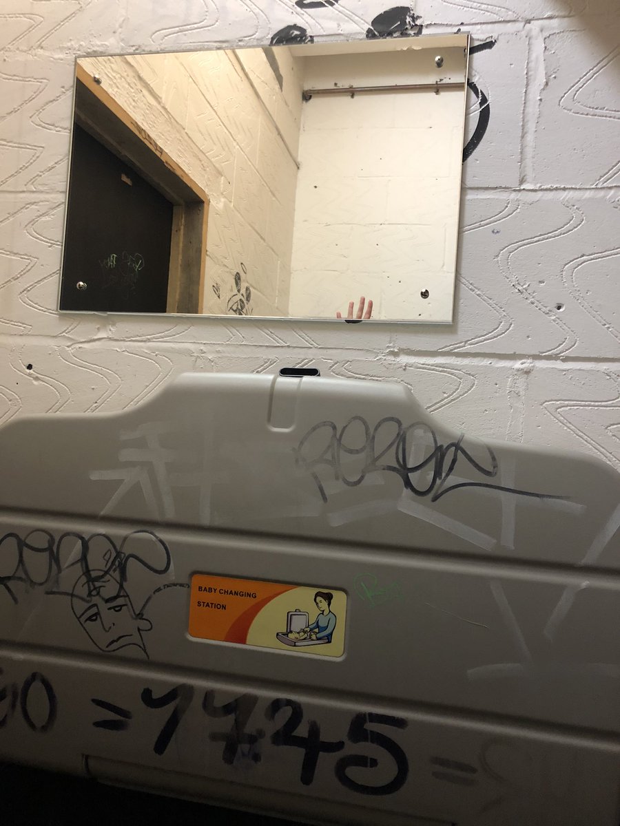 Same photo of A mirror hanging on a bathroom wall where can see a hand waving in the mirror, but in a different bathroom, which this time is covered in graffiti.