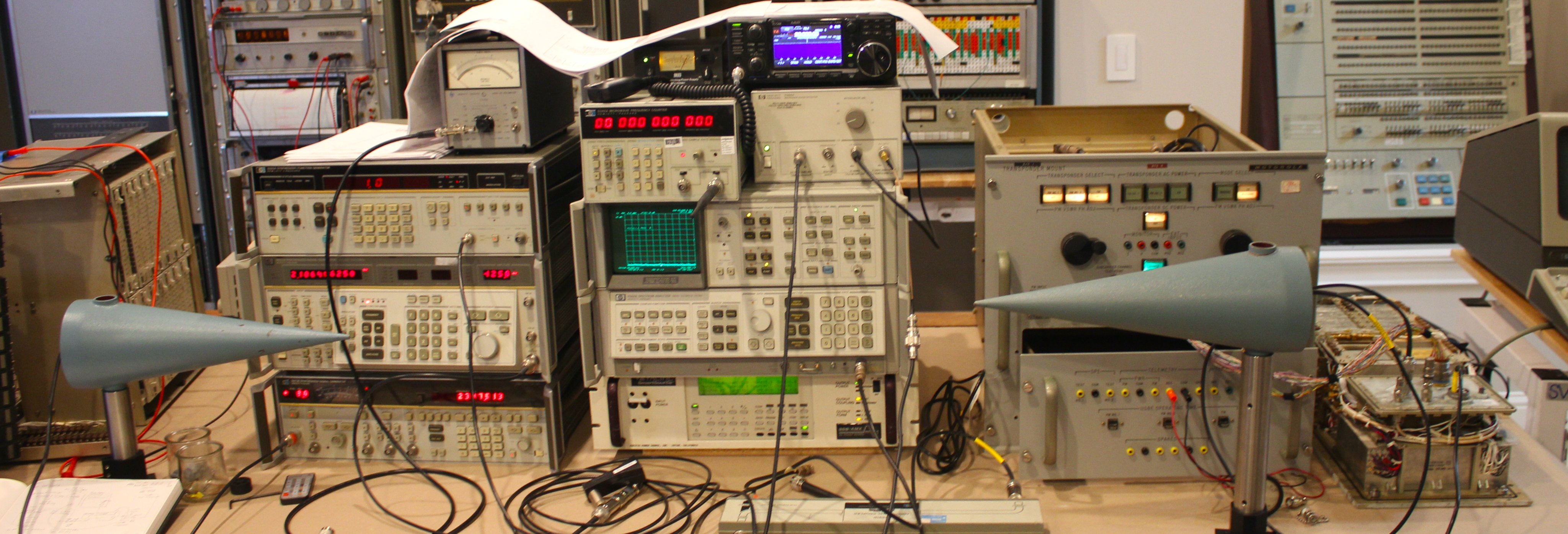 The lab setup to test the S-Band Transponder. In the foreground, two conical antennas are pointed at each other. Three stacks of test equipment are piled up. At the far right is the Transponder. The components are connected with coaxial cables snaking over the desk.