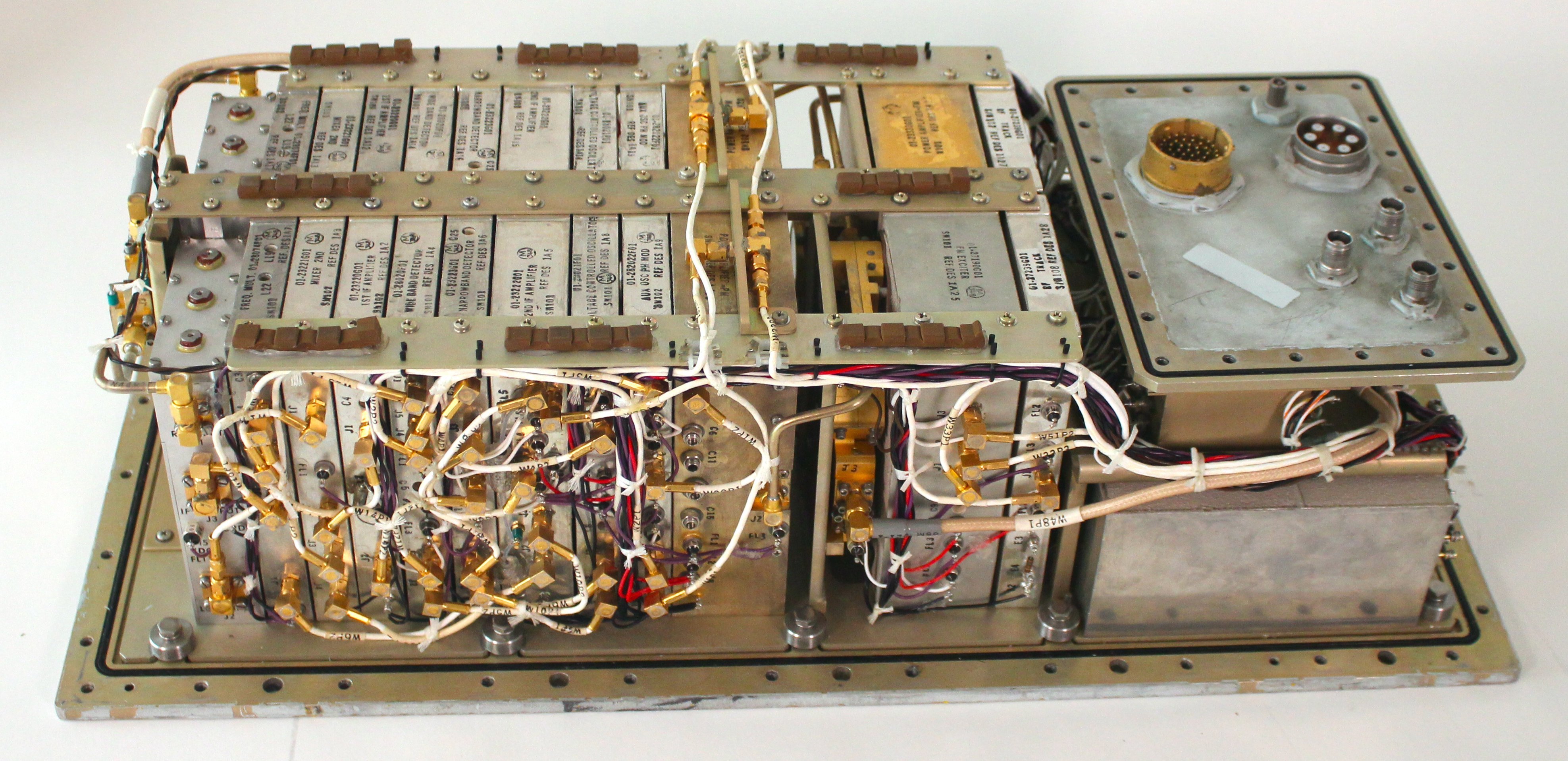 The S-Band Transponder with the cover removed. It is a box with complex wiring of thin coaxial cables, connected to modules with gold connectors. It has military-style round connectors on the top right, along with coaxial connectors.