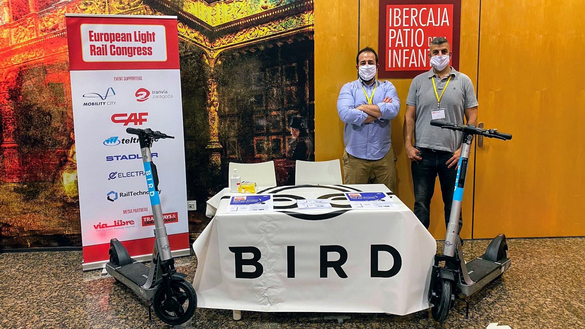 Two men wearing surgical masks stand behind a table with the Bird logo on it. Two Bird scooters are positioned on either side of the table. The setting is indoors. Next to the table there is a sign indicating the European Light Rail Congress. 