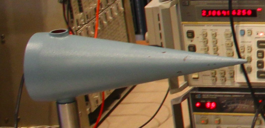 A conical antenna, a bit over a foot long. It is painted greenish-gray. It is in front of the test equipment.